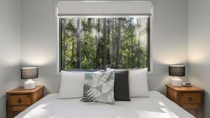 Un dormitorio con un ventanal grande, ropa de cama blanca, dos mesitas de noche con lámparas y vistas a árboles fuera.