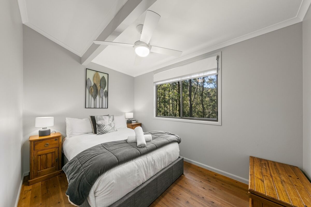 Un dormitorio con cama, mesita de noche, banco de noche, ventilador de techo, ventana con vista a árboles y un pequeño banco de madera.