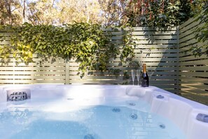 A hot tub with a bottle of champagne and a glass, surrounded by a wooden fence and greenery. - Cottages On Mount View (Millfield)