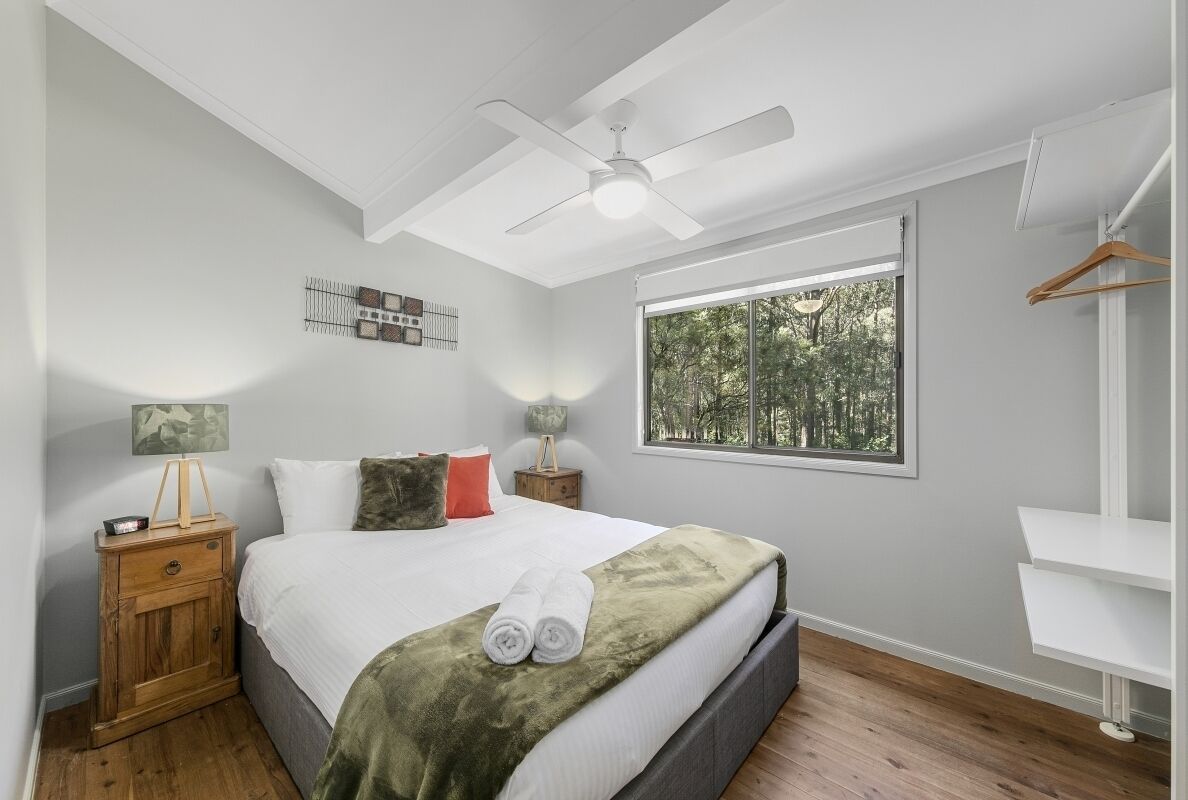 Quarto com cama, ventilador de teto, janela com vista para árvores, mesa de cabeceira de madeira e guarda-roupa branco.
