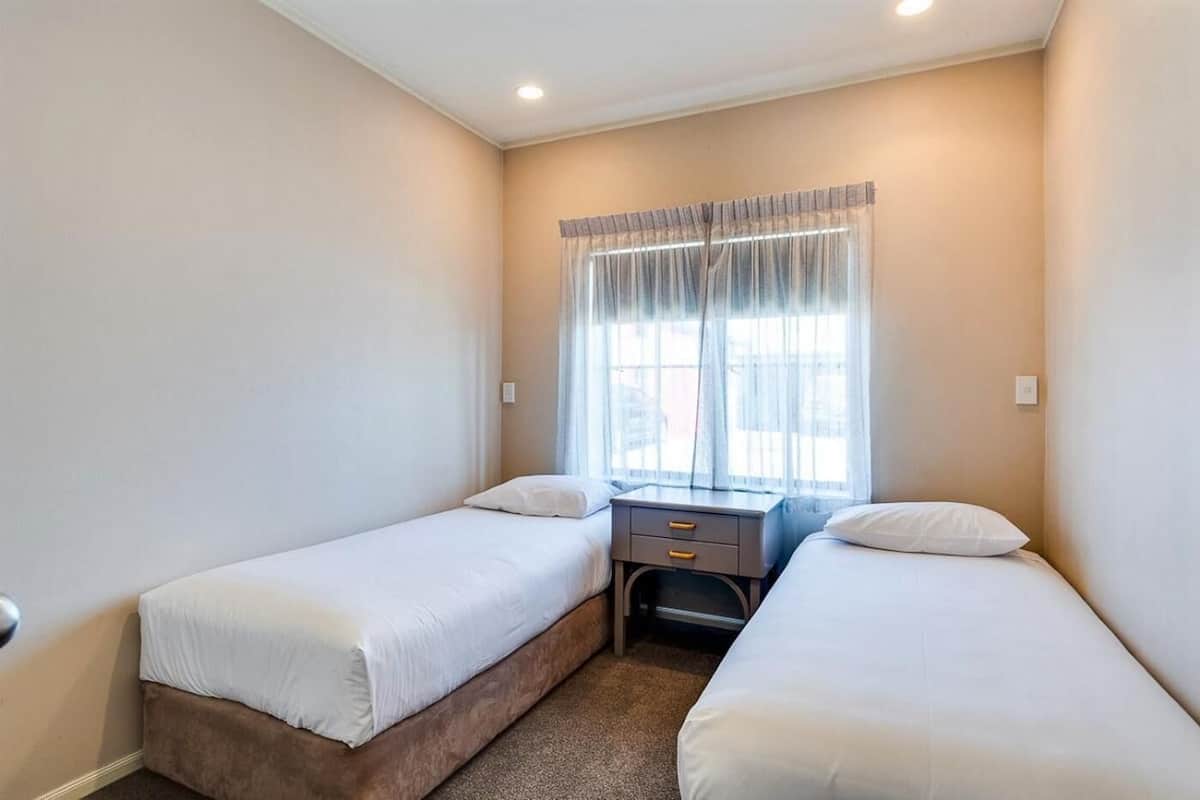 A small, clean room with two single beds, a nightstand, and a window with sheer curtains.