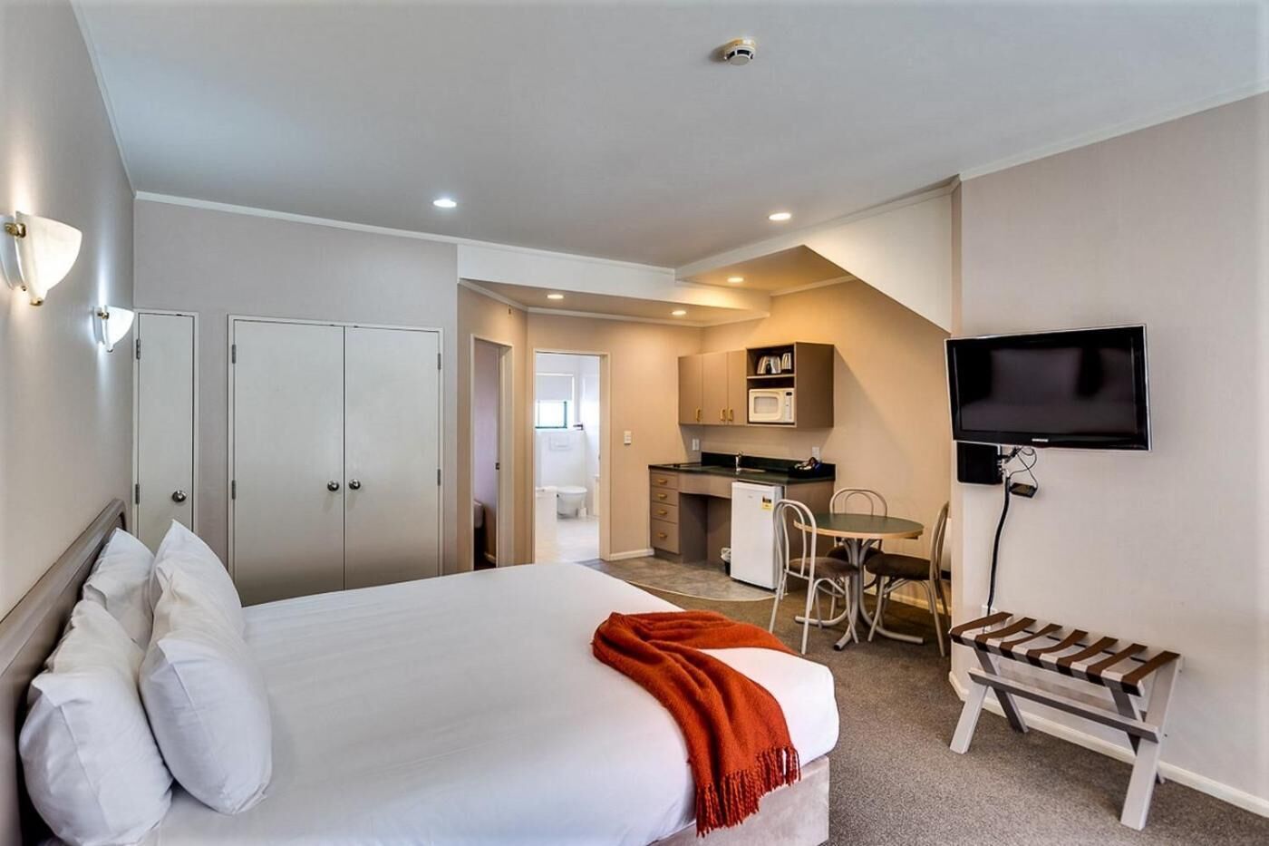 A hotel room with a bed, a desk, a chair, a television, and a kitchenette.