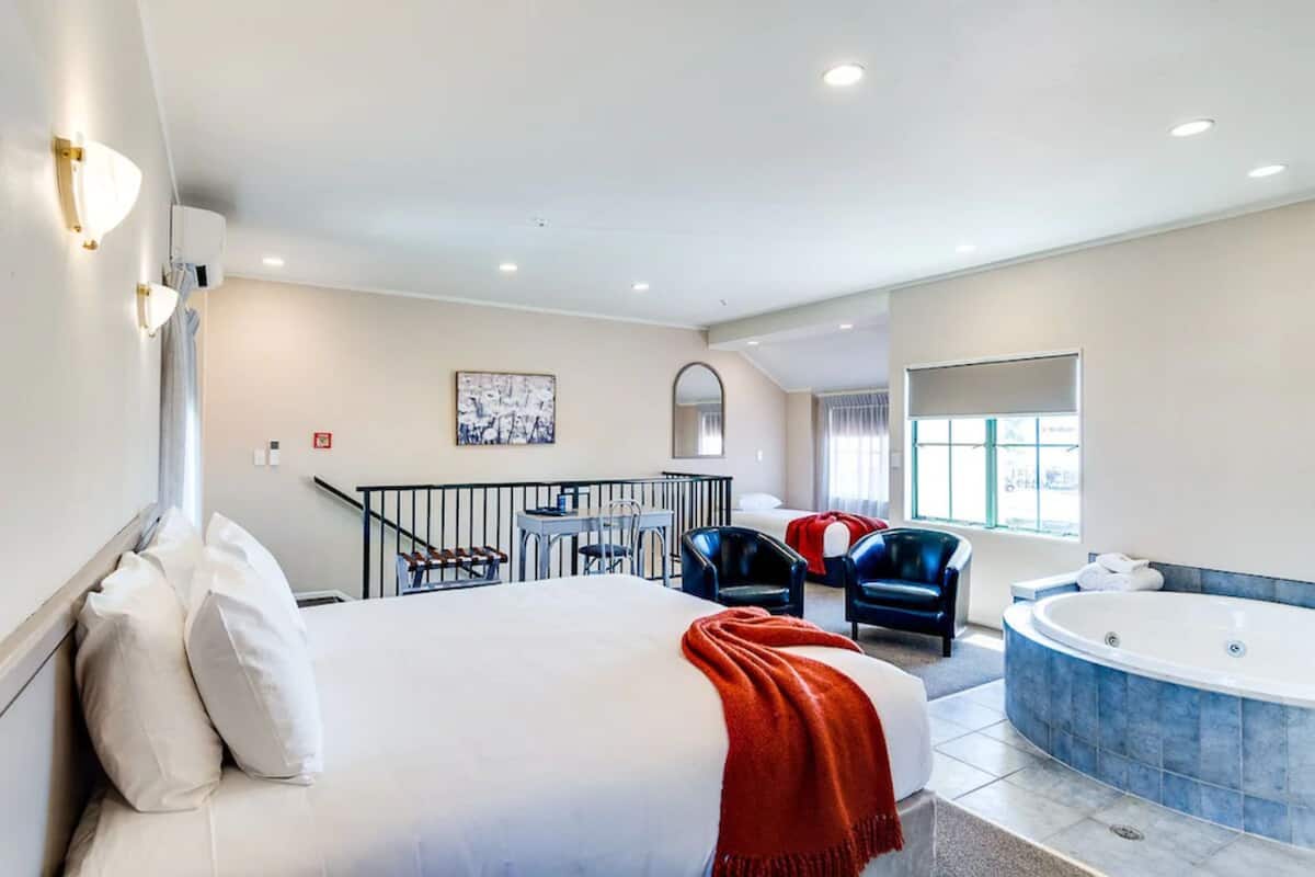 A hotel room with a large bed, a sitting area with chairs, a small table, a balcony, and a jacuzzi.