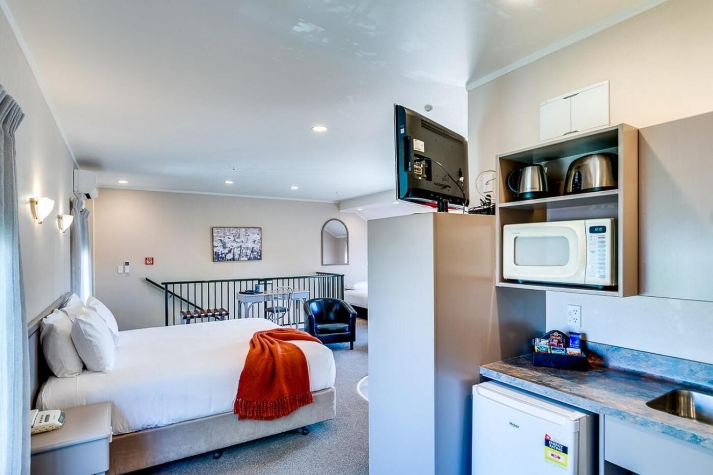 A compact hotel room with a bed, a small kitchenette, a flat-screen TV, and a view of the outside.