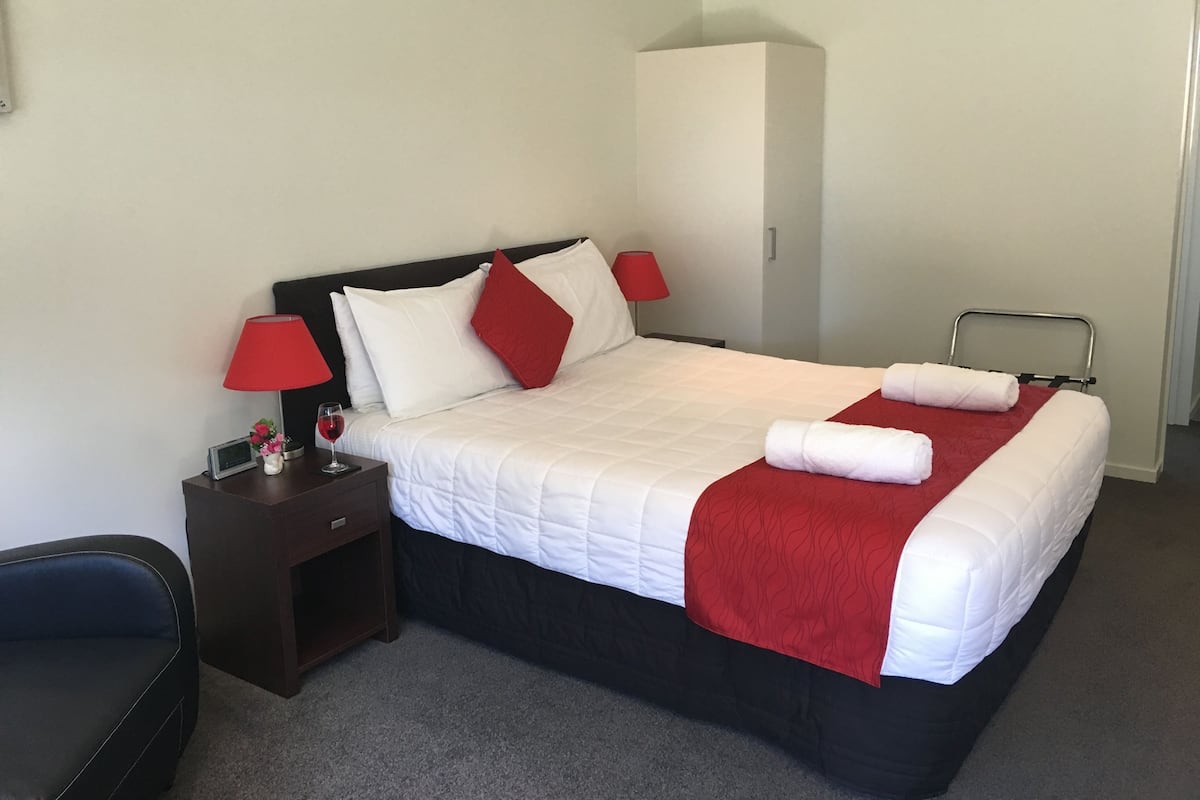 A neatly made bed with white and red bedding, a red lamp, a small table with a digital clock, and a dark-colored chair.