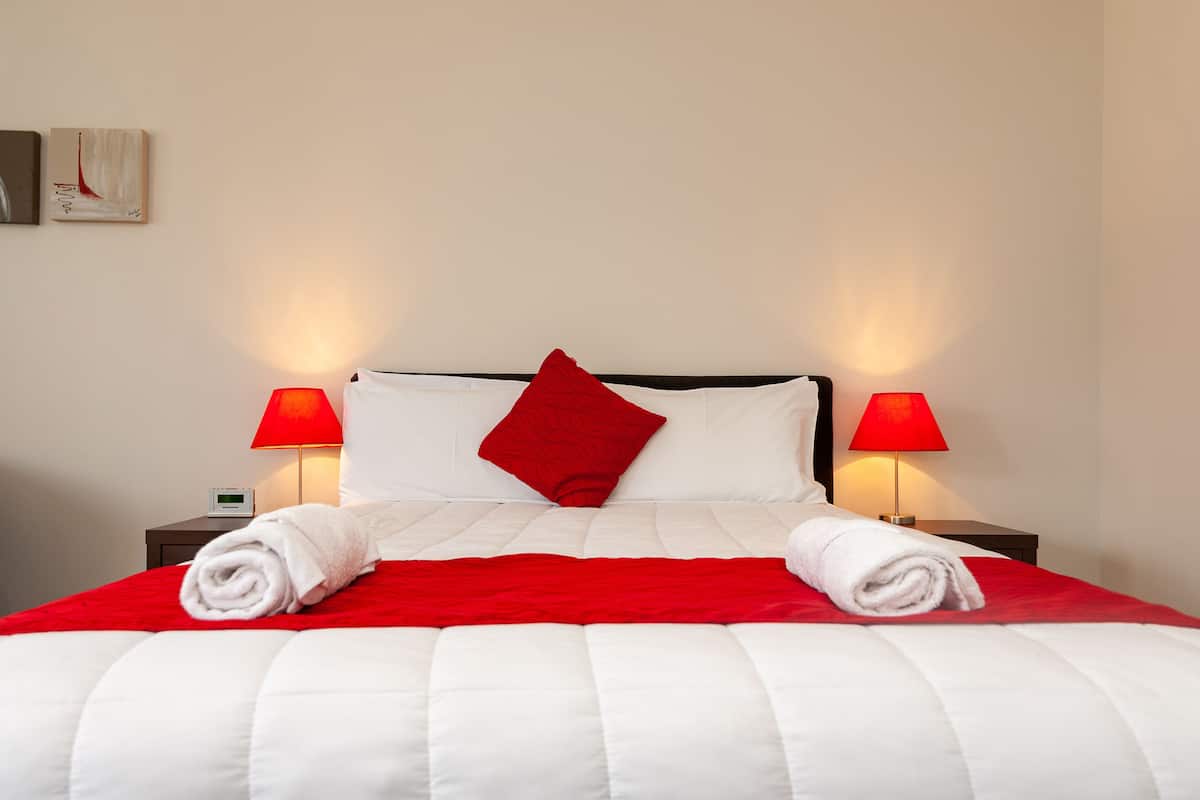 A neatly made bed with white and red bedding, two bedside lamps, and a digital clock on a bedside table.
