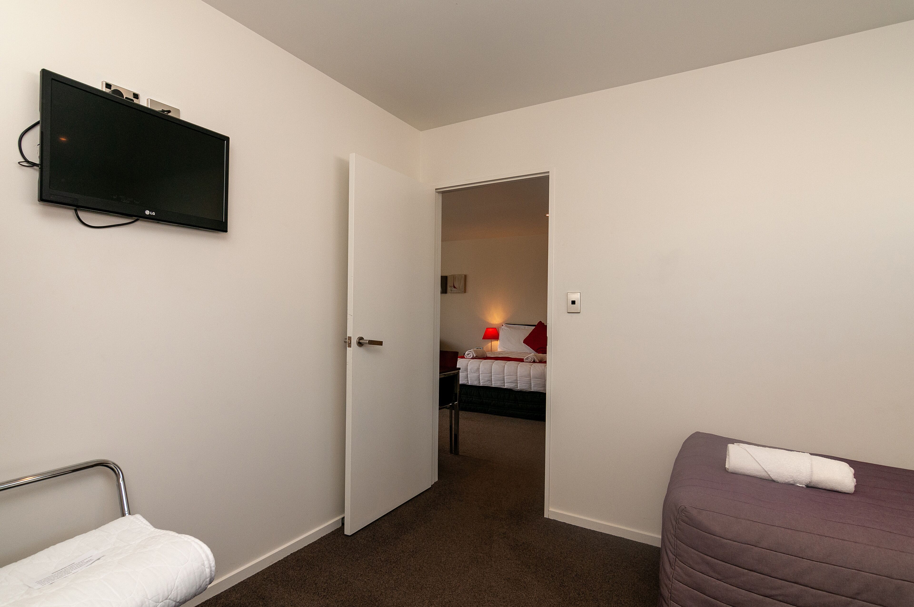 Una habitación de hotel con una cama, una mesita de noche, un televisor montado en la pared y una puerta que da a otra habitación.