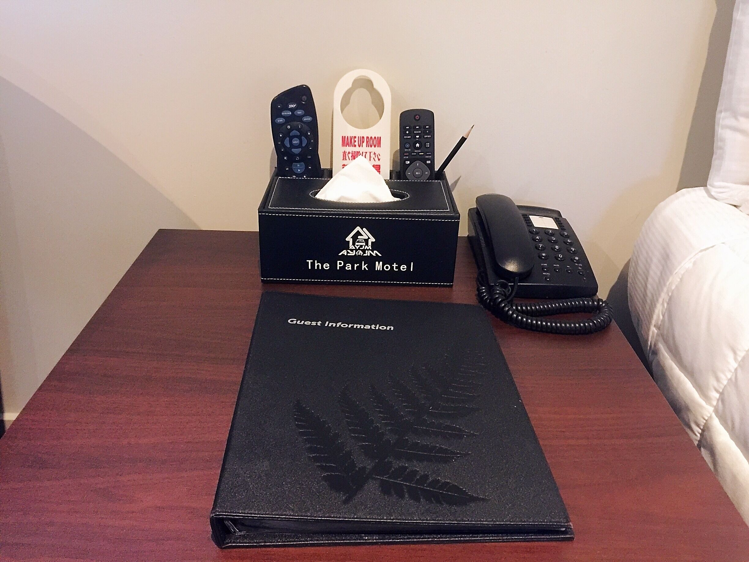 Habitación de hotel con una mesita de noche de madera que contiene un folleto informativo para huéspedes, un teléfono negro y un estuche con mandos a distancia y pañuelos de papel.