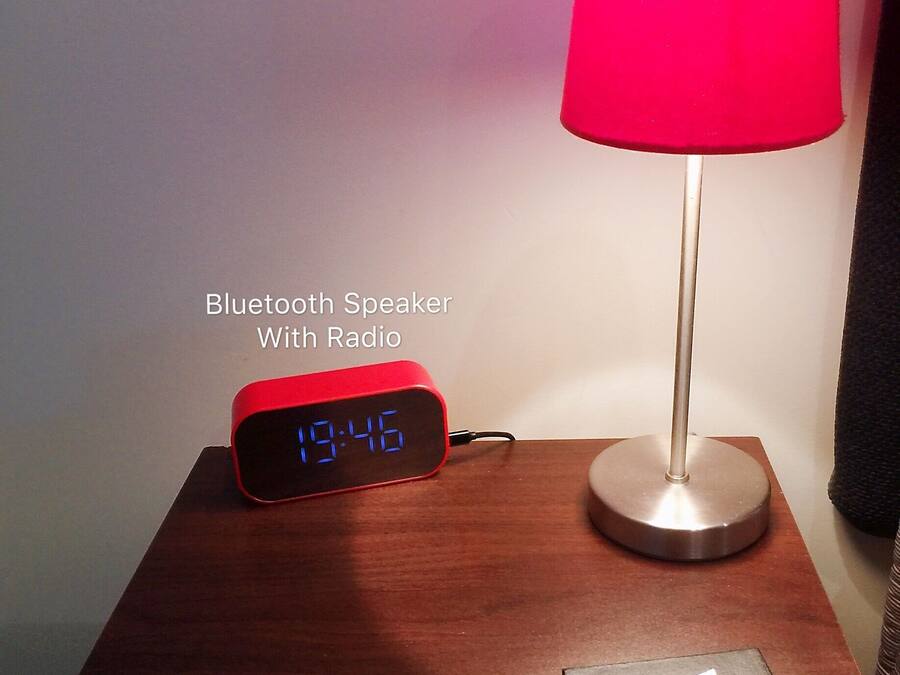 A Bluetooth speaker with a digital display on a wooden table, next to a red lamp and a sign for 'The Park Motel'.