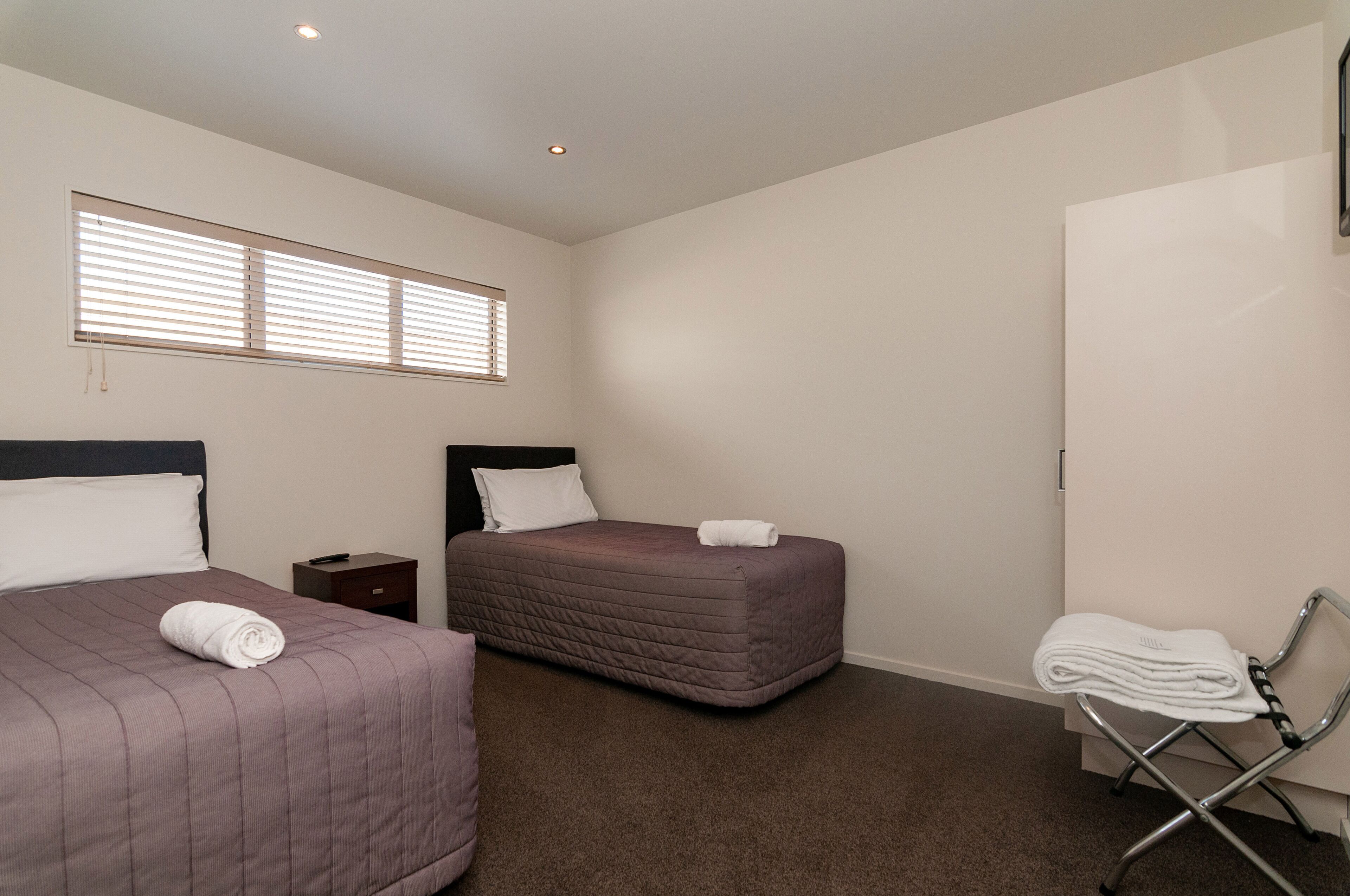 Quarto de hotel com duas camas, uma mesinha de cabeceira, uma cadeira e uma janela com persianas.