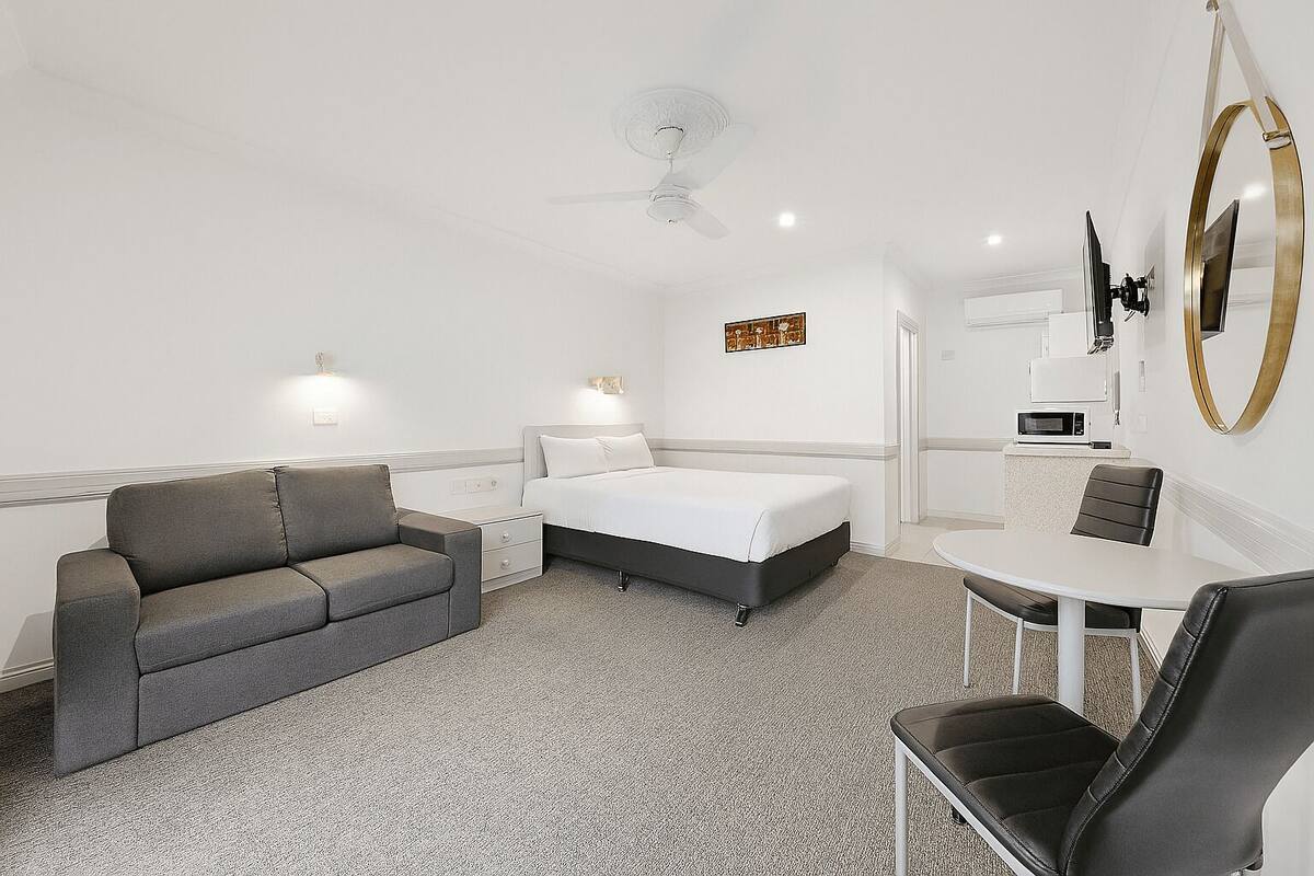 Habitación de hotel con una cama, un sofá gris, una mesita con una silla, ventilador de techo, microondas y un espejo grande.