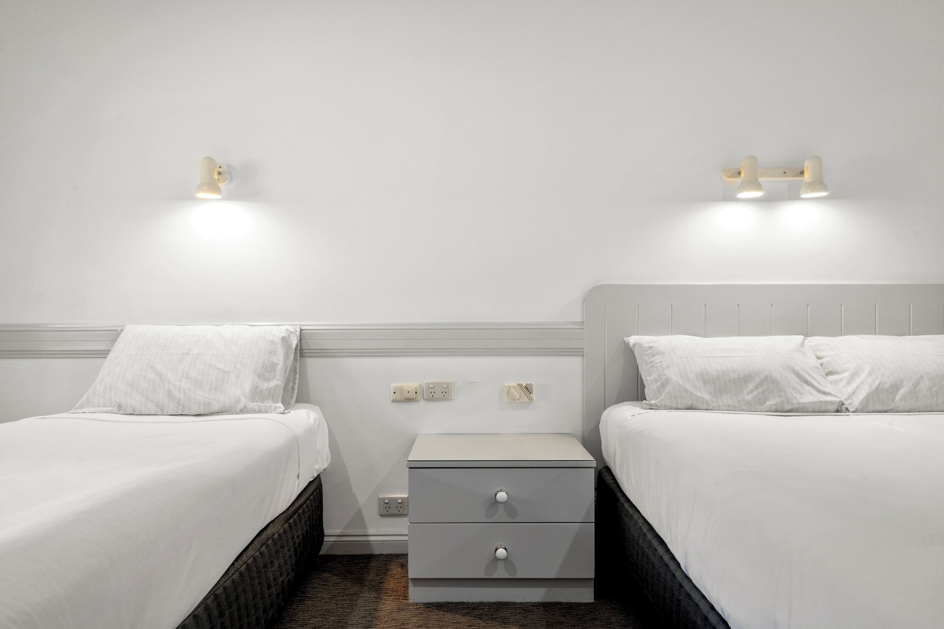 Et hotelværelse med to senge, et lille natbord med skuffer og vægmonteret belysning.
