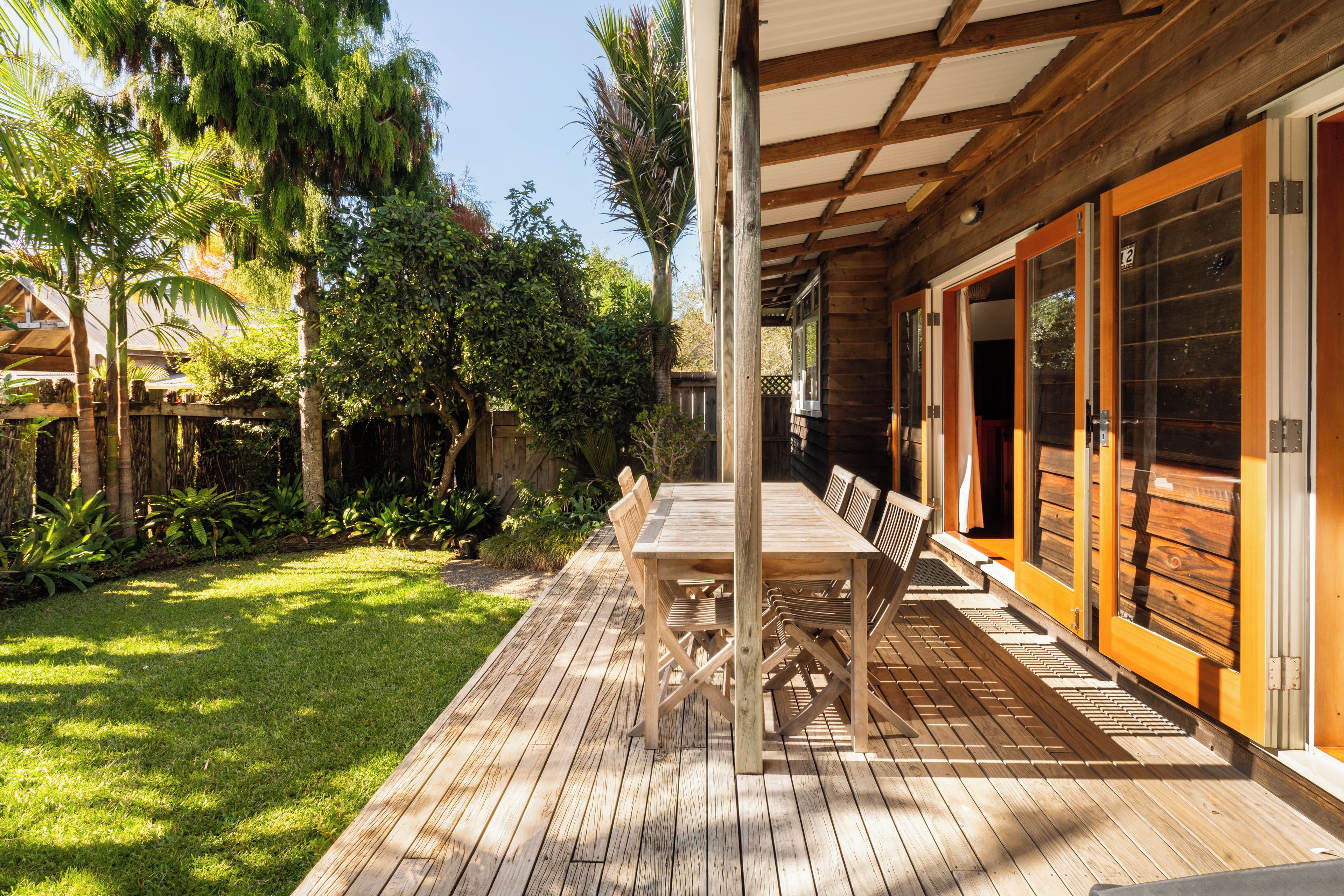 A wooden deck with a table and chairs, surrounded by lush greenery and a wooden building with large windows.