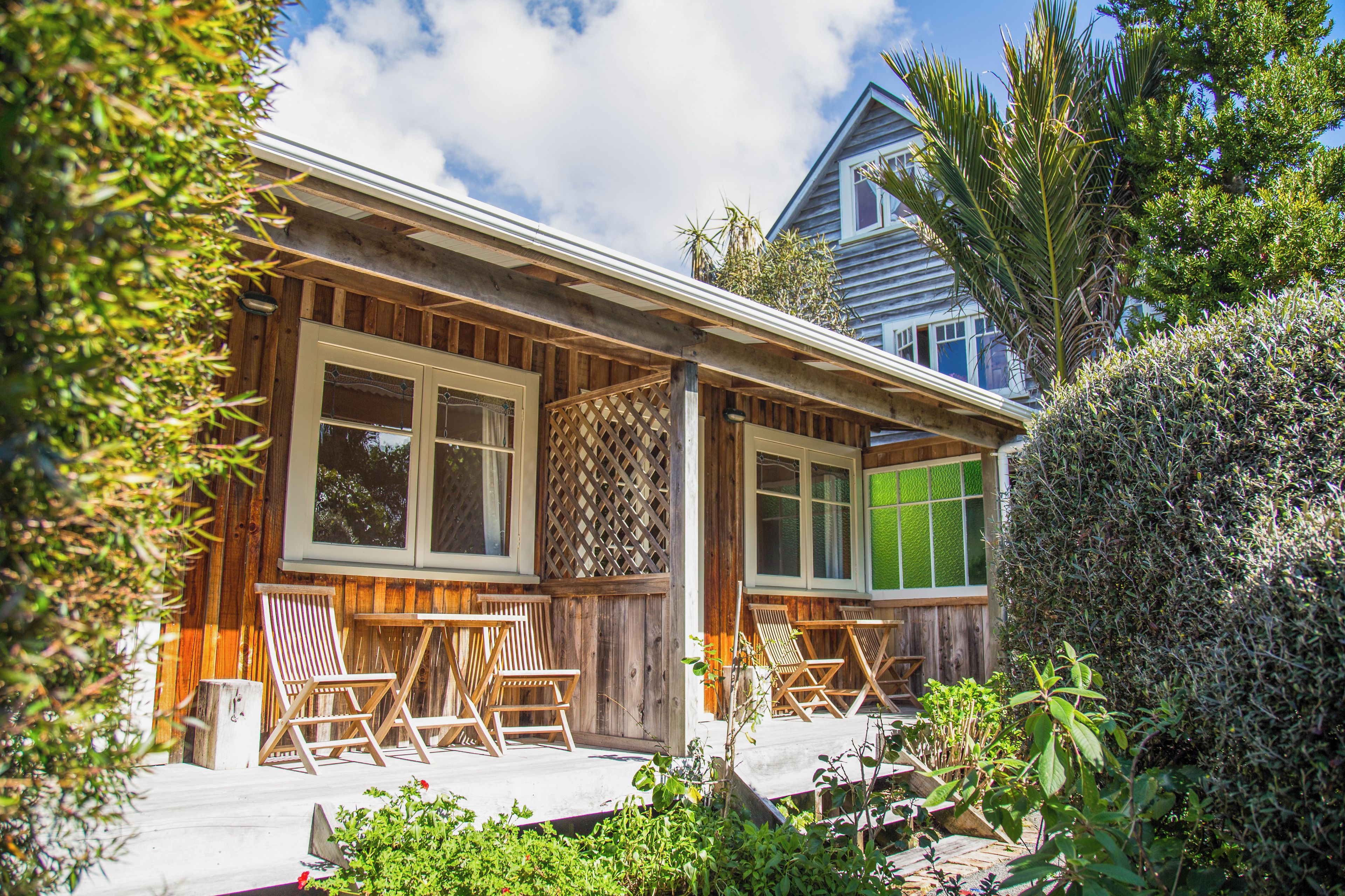 A wooden house with a porch, two windows, and a small garden area with chairs.