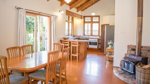 A spacious kitchen with wooden cabinets and a large island, a dining area with wooden chairs, and a stone fireplace. - Church Accommodation (Hahei)