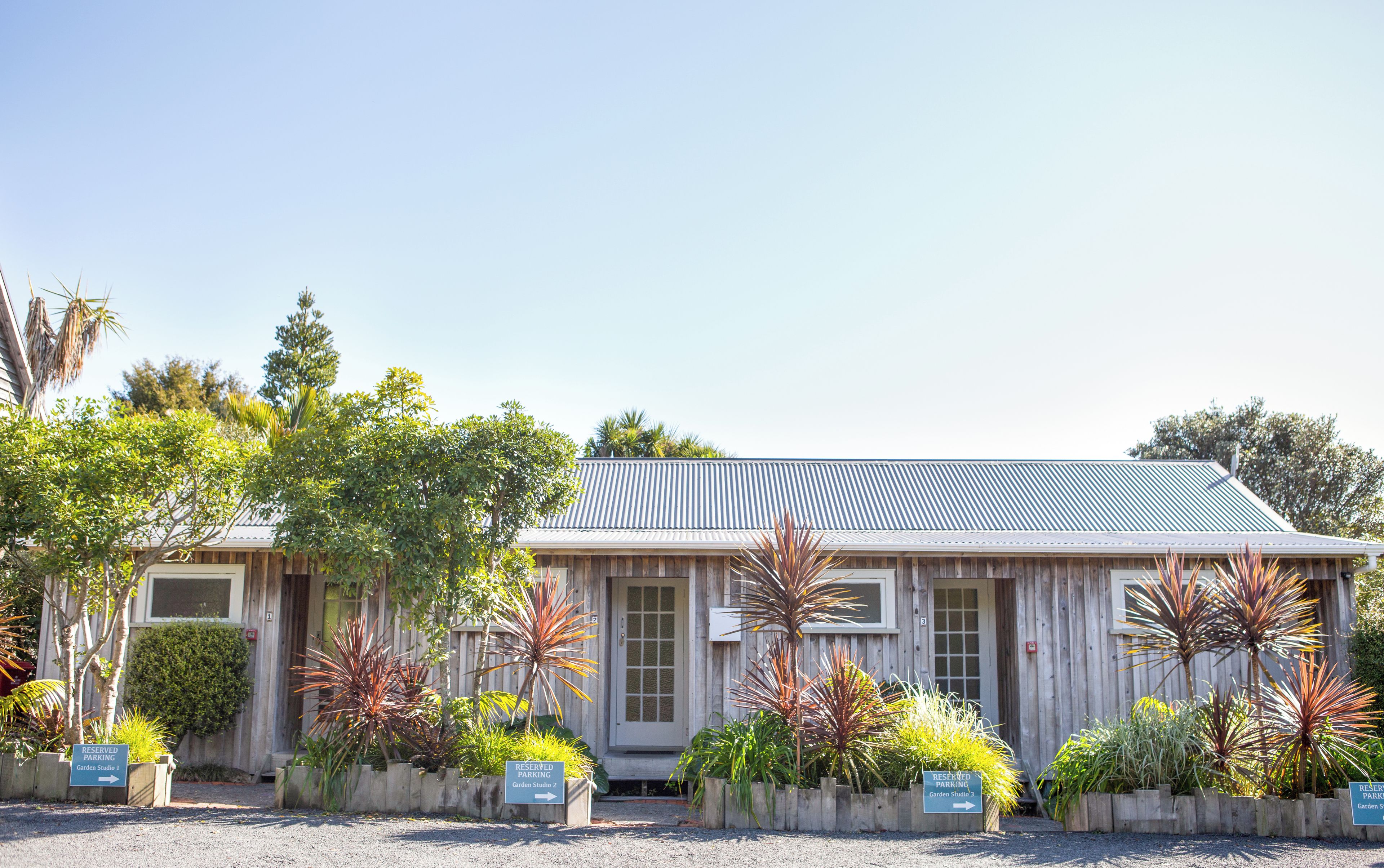 A building with a corrugated metal roof, wooden walls, and multiple entry points. There are signs indicating reserved parking for Garden Studios 1, 2, and 3.