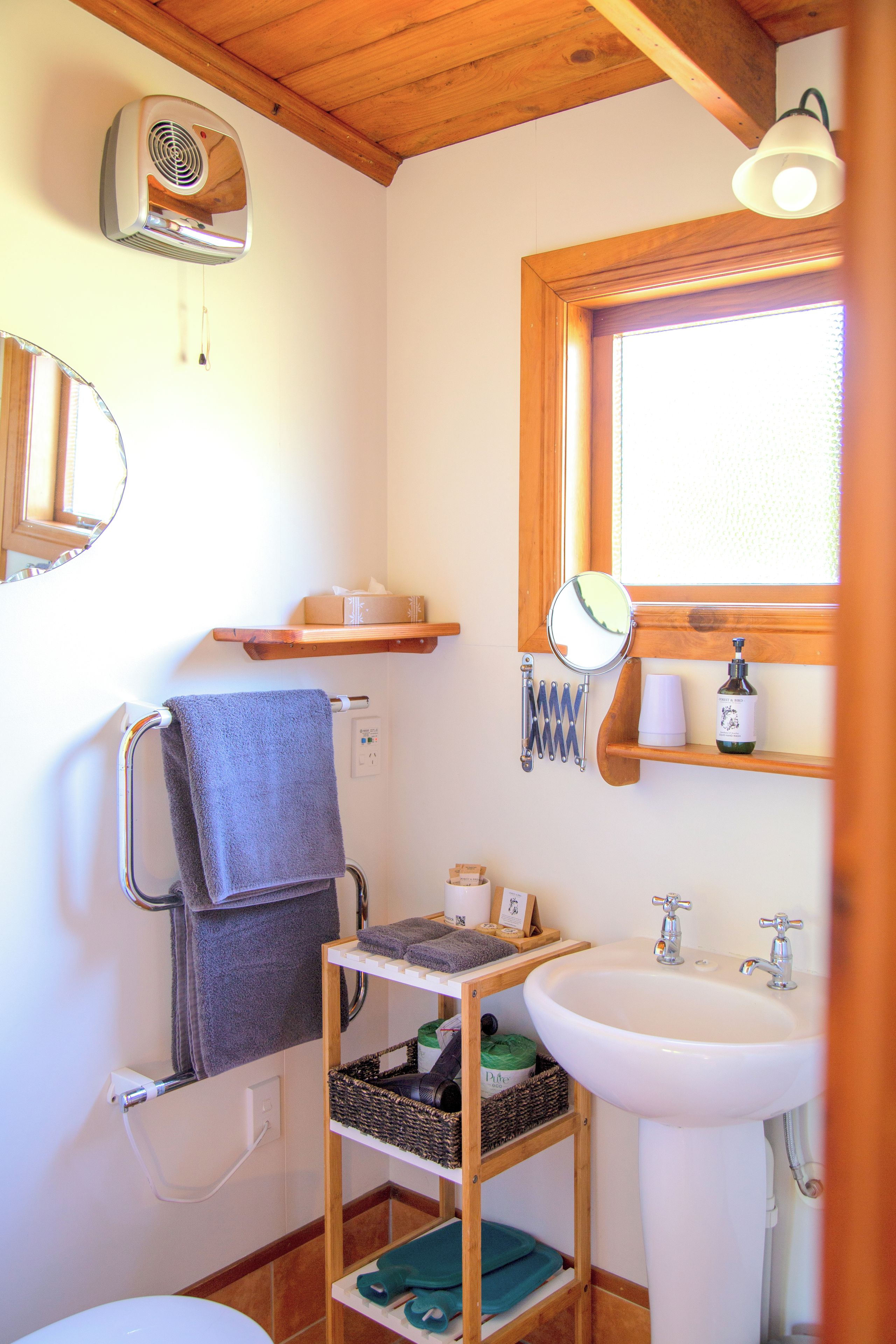 Een badkamer met een witte wasbak, een spiegel, een handdoekhouder, een plank met toiletartikelen en een raam met zonwering.