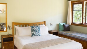 A bedroom with a wooden bed, a nightstand, a bedside lamp, a window with a view, and a wooden ceiling. - Church Accommodation (Hahei)