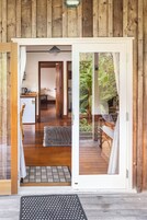 Puerta abierta que da a un interior de madera con una zona de cocina, una mesa de comedor y vistas a un jardín a través de una puerta de cristal.