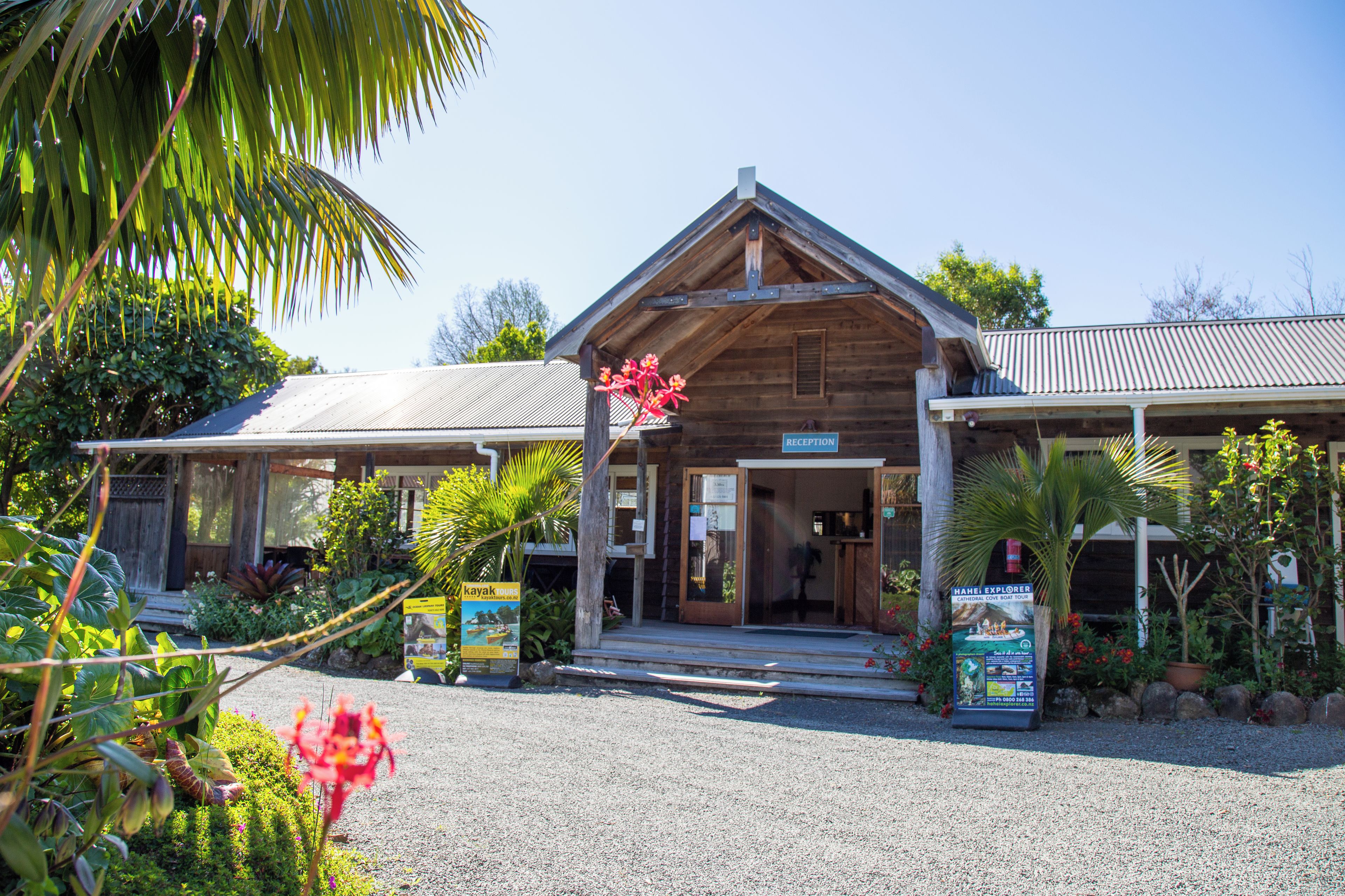 A wooden building with a covered entrance, surrounded by tropical plants and a gravel driveway.