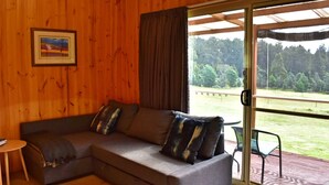 Una habitación acogedora con un sofá seccional, una mesita y un amplio ventanal corredera que da a un espacio exterior con vistas a árboles y un campo cubierto de césped.