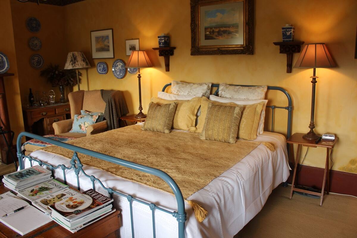 A neatly made bed with a metal frame, a bedside table with books and a lamp, and a wall with framed pictures and decorative plates.
