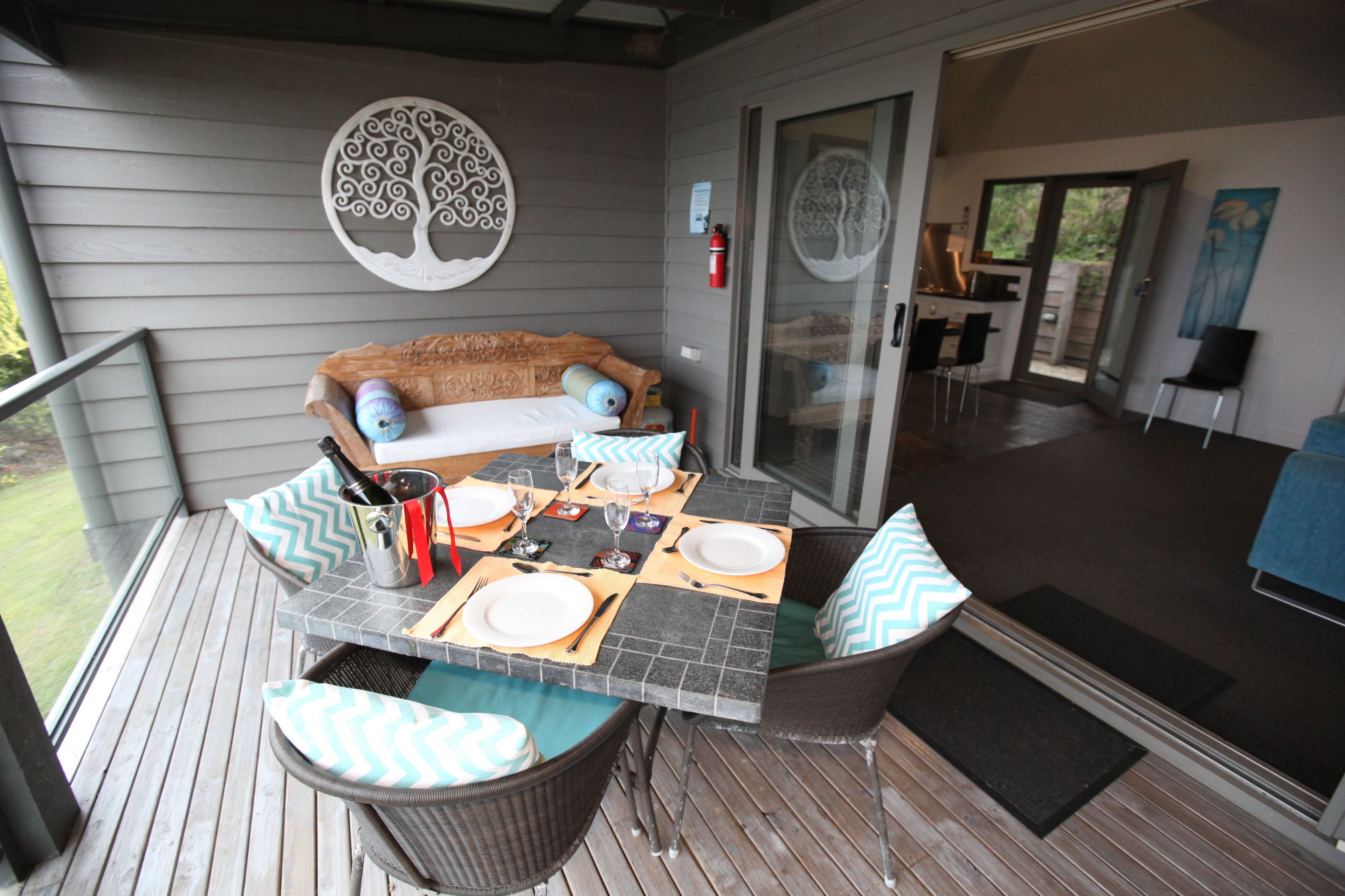 A covered outdoor dining area with a table set for two, a cushioned bench, and a decorative wall piece.