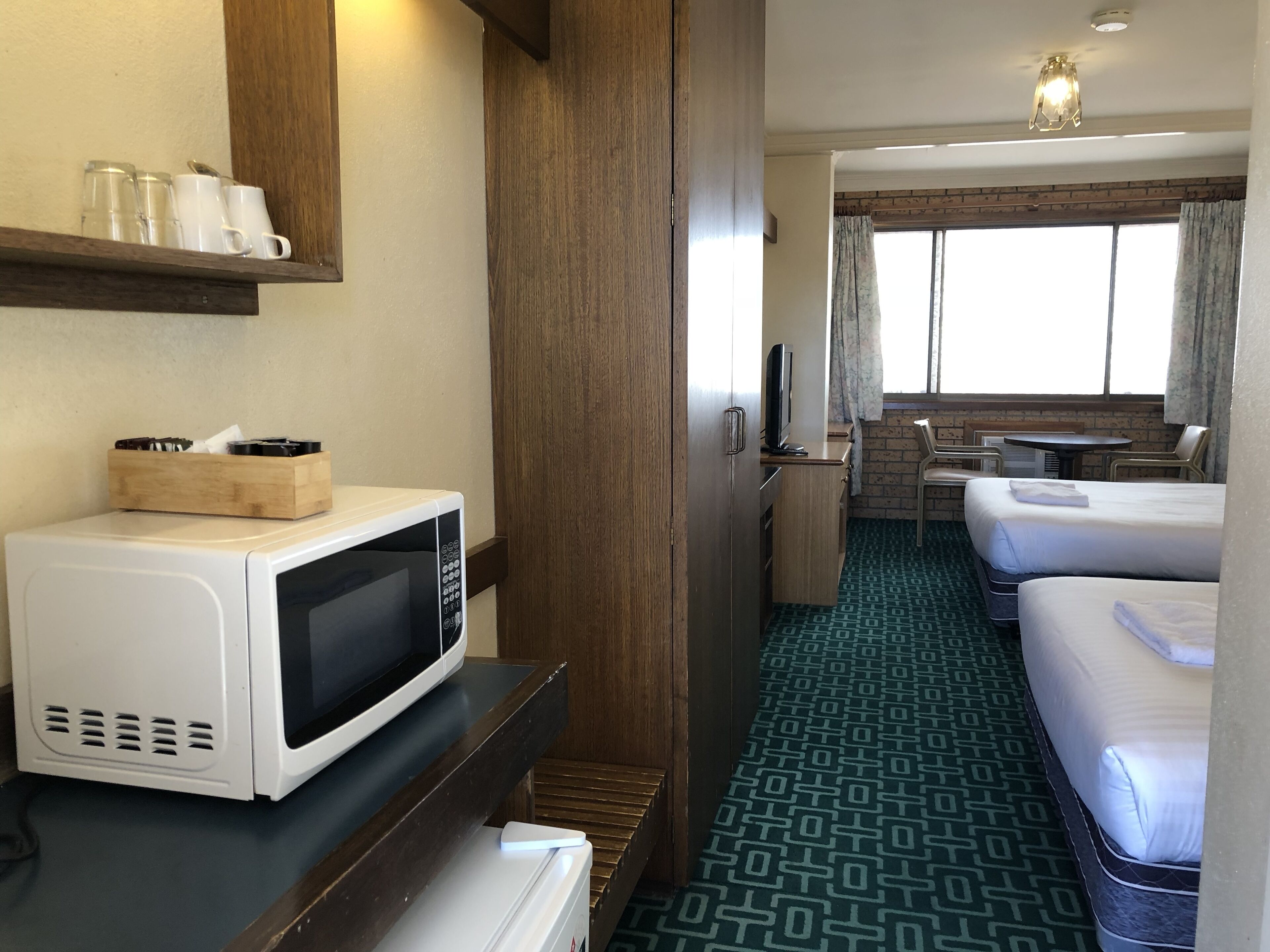 A compact hotel room with a kitchenette, two beds, a small table, and a window with curtains.