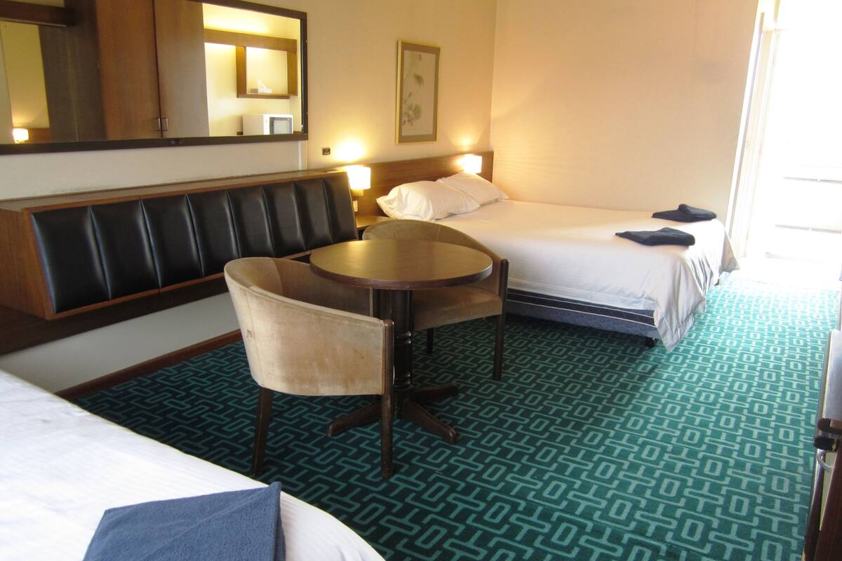 Ein Hotelzimmer mit zwei Betten, einer Sitzecke mit Tisch und Stühlen, einem großen Spiegel und einem gemusterten Teppich.
