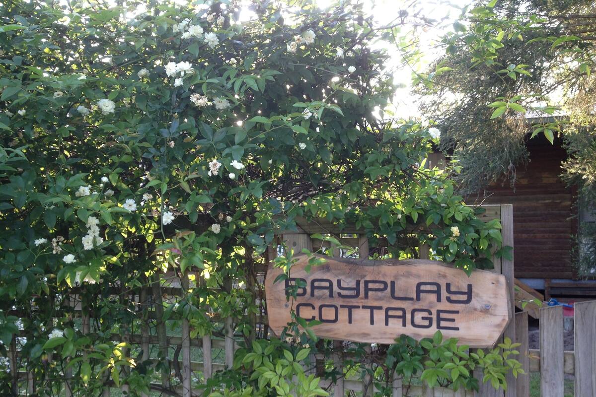 Une pancarte en bois portant l’inscription « Bayplay Cottage », avec en arrière-plan une végétation luxuriante et une clôture en bois.