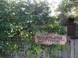 A wooden sign with the text "Bayplay Cottage" against a backdrop of lush greenery and a wooden fence. - Bayplay Beach Accommodation - 2 locations (Portsea)