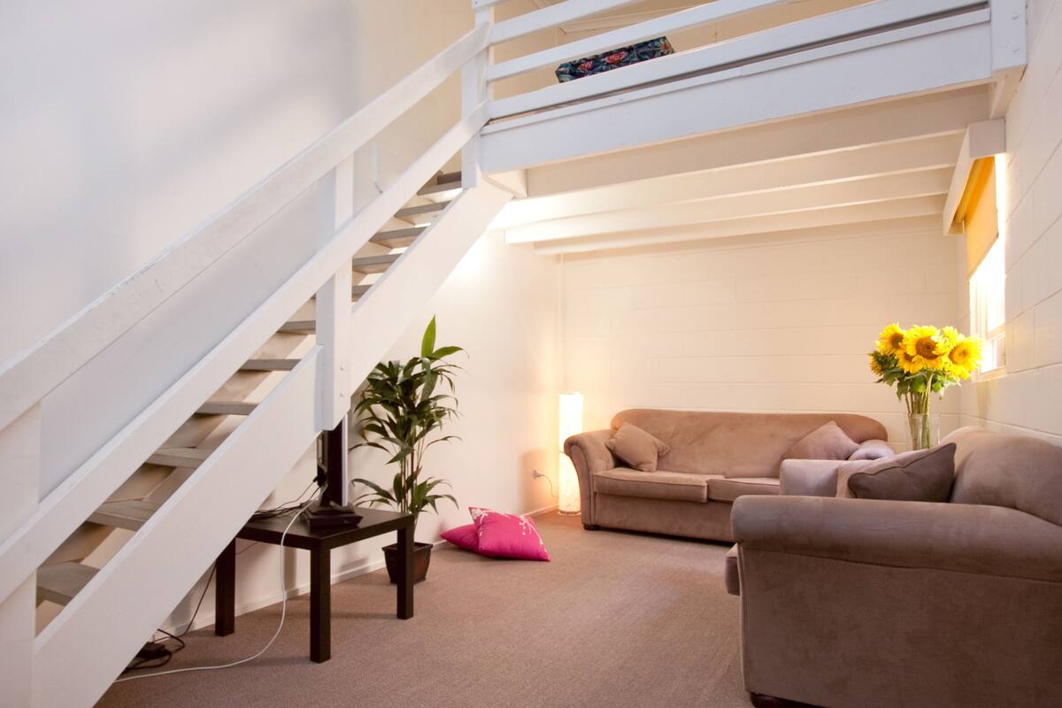 Un salon avec un escalier, un canapé, une table basse, une plante en pot et un vase rempli de fleurs.
