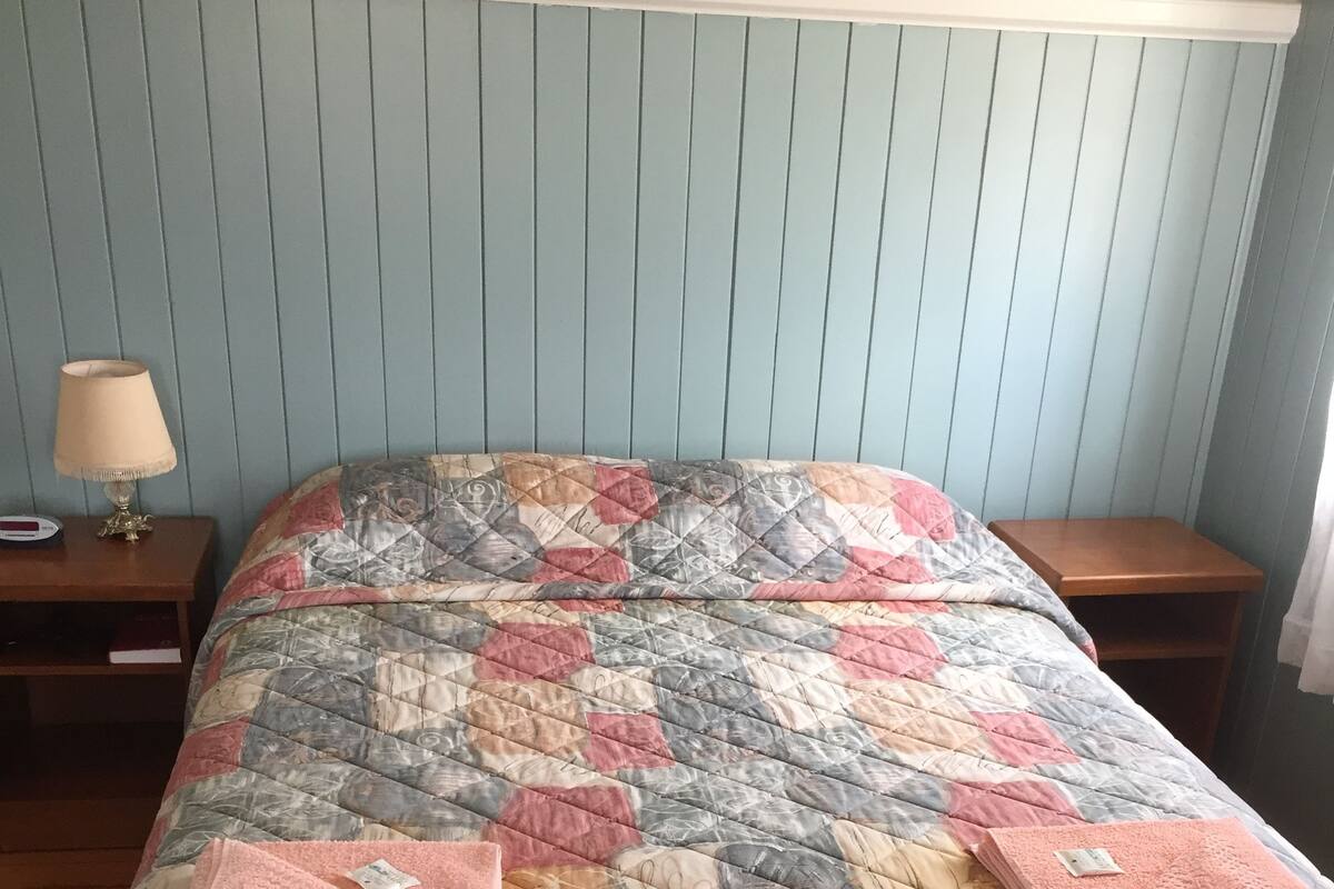 A bedroom with a quilted bedspread, two wooden nightstands, a bedside lamp, and a window with curtains.