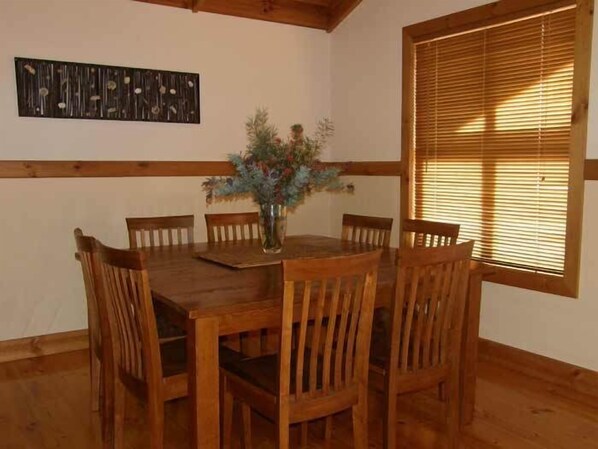 A dining area with a wooden table and chairs, a vase with flowers, and a window with blinds.