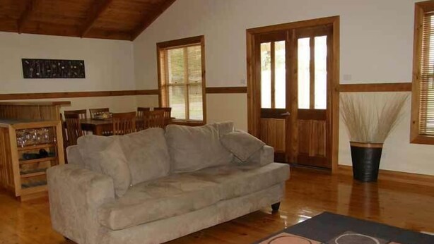 Un salón con un sofá beige, suelo de madera, una zona de comedor con sillas y una mesa, y un ventanal con persianas.
