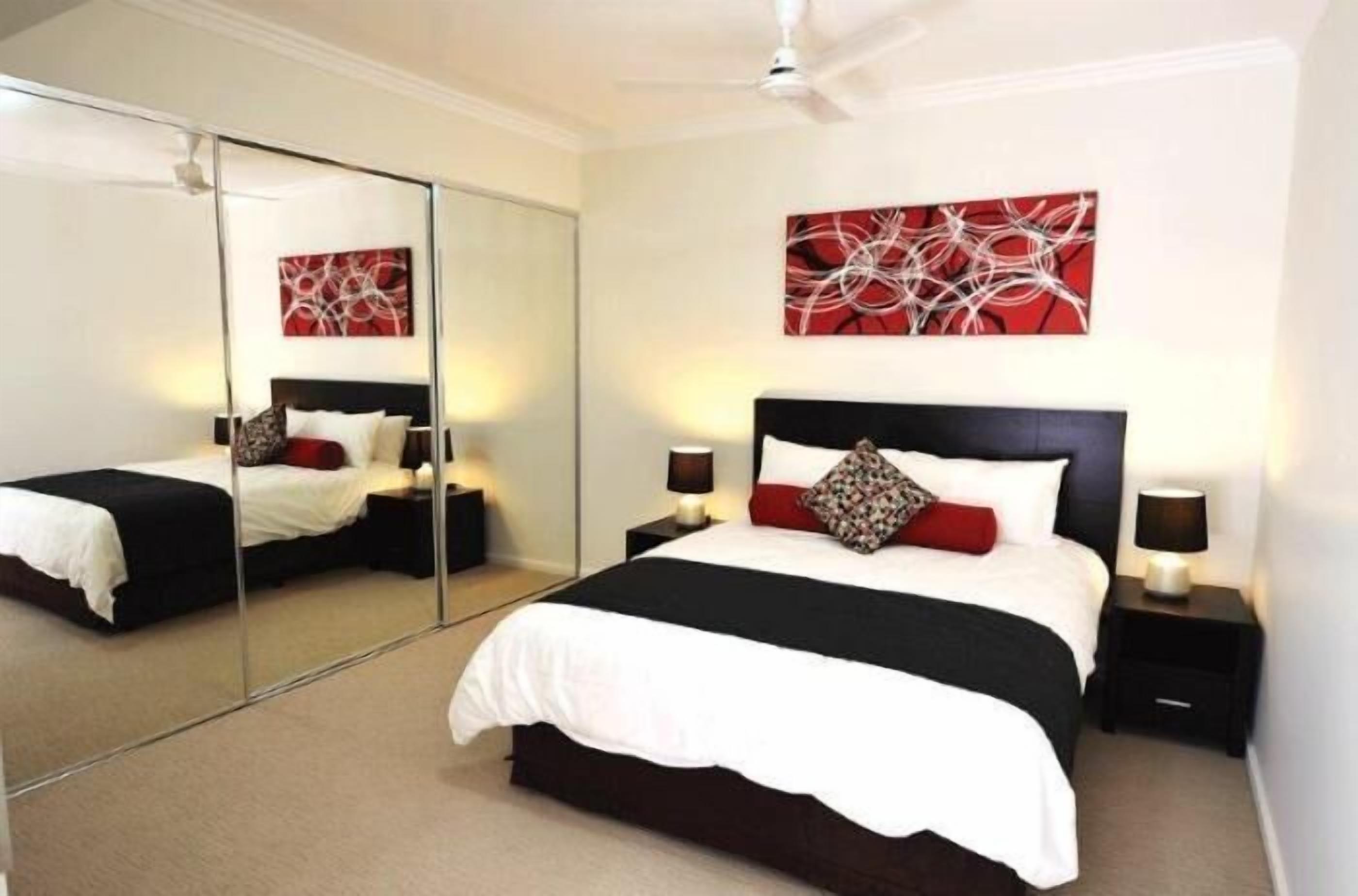 A modern bedroom with a large mirror, a bed with black and white bedding, two bedside tables with lamps, and abstract red and white artwork on the wall.