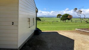 A small, single-story building with a number '2' on its side, situated in a rural area with a clear sky and a grassy field in the background. - Promhills Cabins (Yanakie)