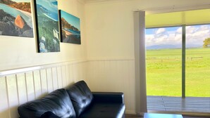 A living room with a black leather sofa, a wooden coffee table, and a large window offering a view of a green field and a distant mountain range. - Promhills Cabins (Yanakie)