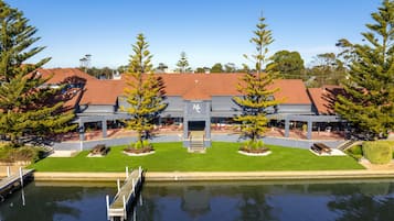 A modern building with a red roof, surrounded by a water body, and two large trees in front.