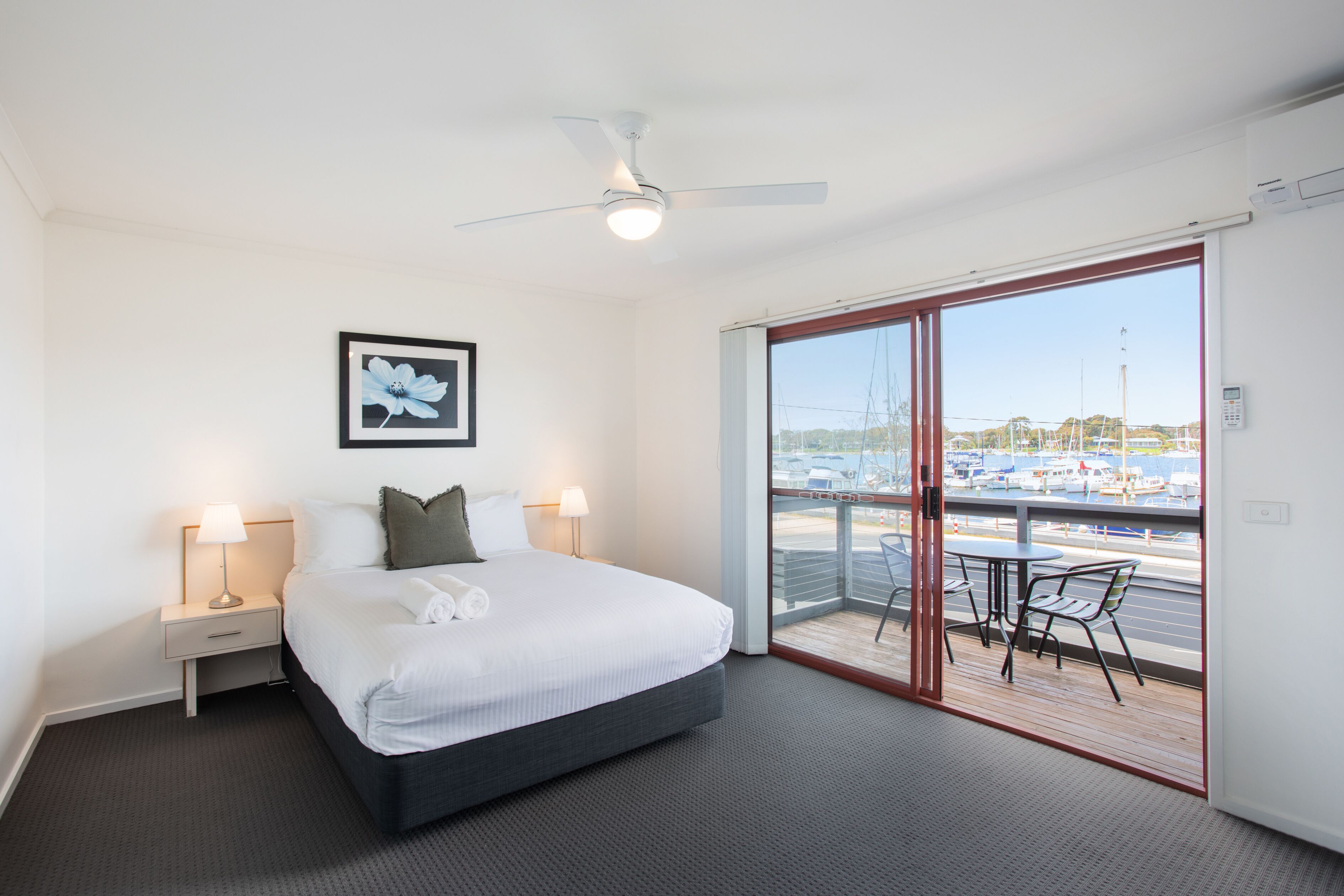 Habitación de hotel con cama, lámparas de noche, ventilador de techo, ventana grande con vistas a barcos y balcón con mesa y sillas.