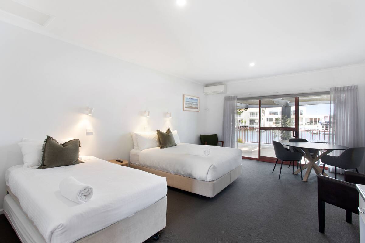 Habitación de hotel con dos camas, un balcón con mesa y sillas, y vistas a la ciudad.
