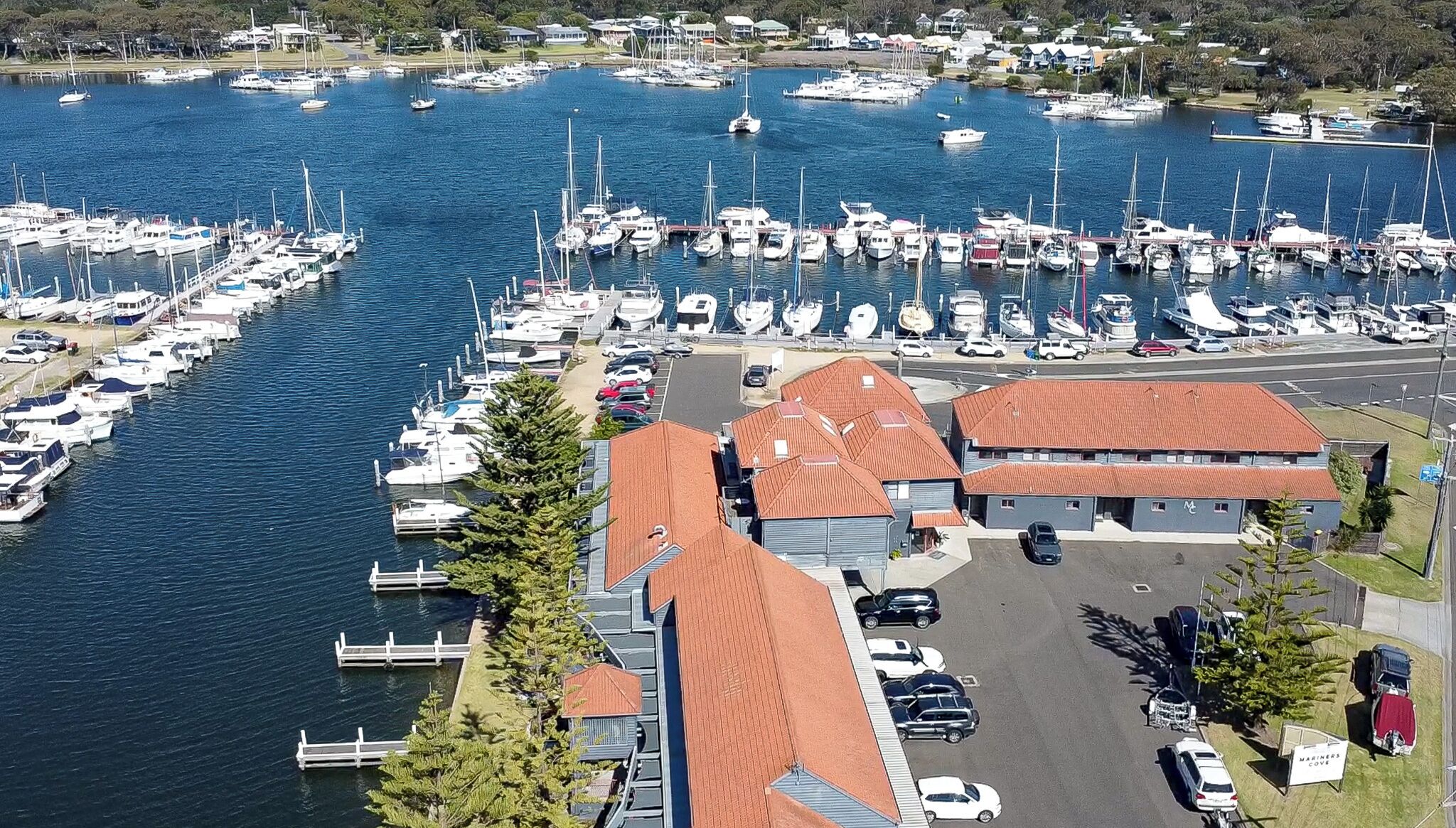 En småbåtshamn med många båtar förtöjda, en parkeringsplats och en byggnad med ett rött tak.