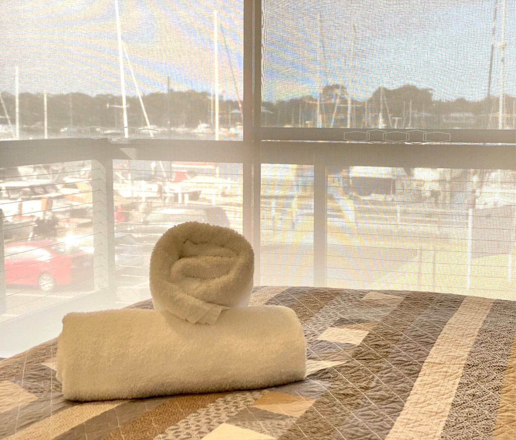 Een keurig opgevouwen witte handdoek op een bed met een decoratieve spreideken, met uitzicht op een jachthaven door een groot raam.