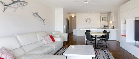 Una sala de estar moderna con un sofá blanco, una mesa de centro y un comedor con una mesa y sillas. Se observa una cocina al fondo con armarios blancos y un microondas.