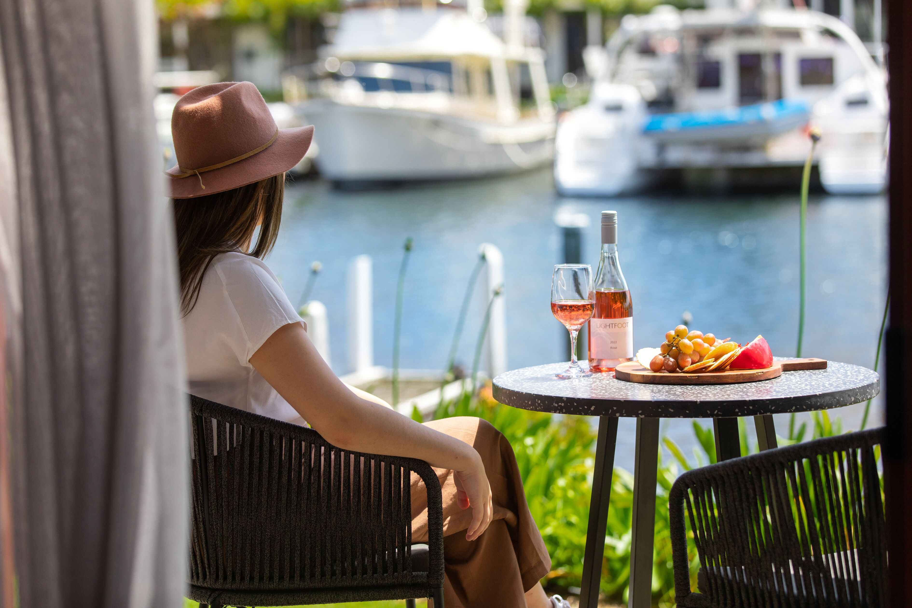 Une personne est assise à une table ronde, devant une bouteille de vin, un verre et une assiette de raisins et de pastèque. La table est placée à l’extérieur, avec vue sur des bateaux sur l’eau.