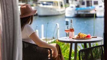 A person seated at a round table with a bottle of wine, a glass, and a plate of grapes and watermelon. The table is outdoors with a view of boats on the water.