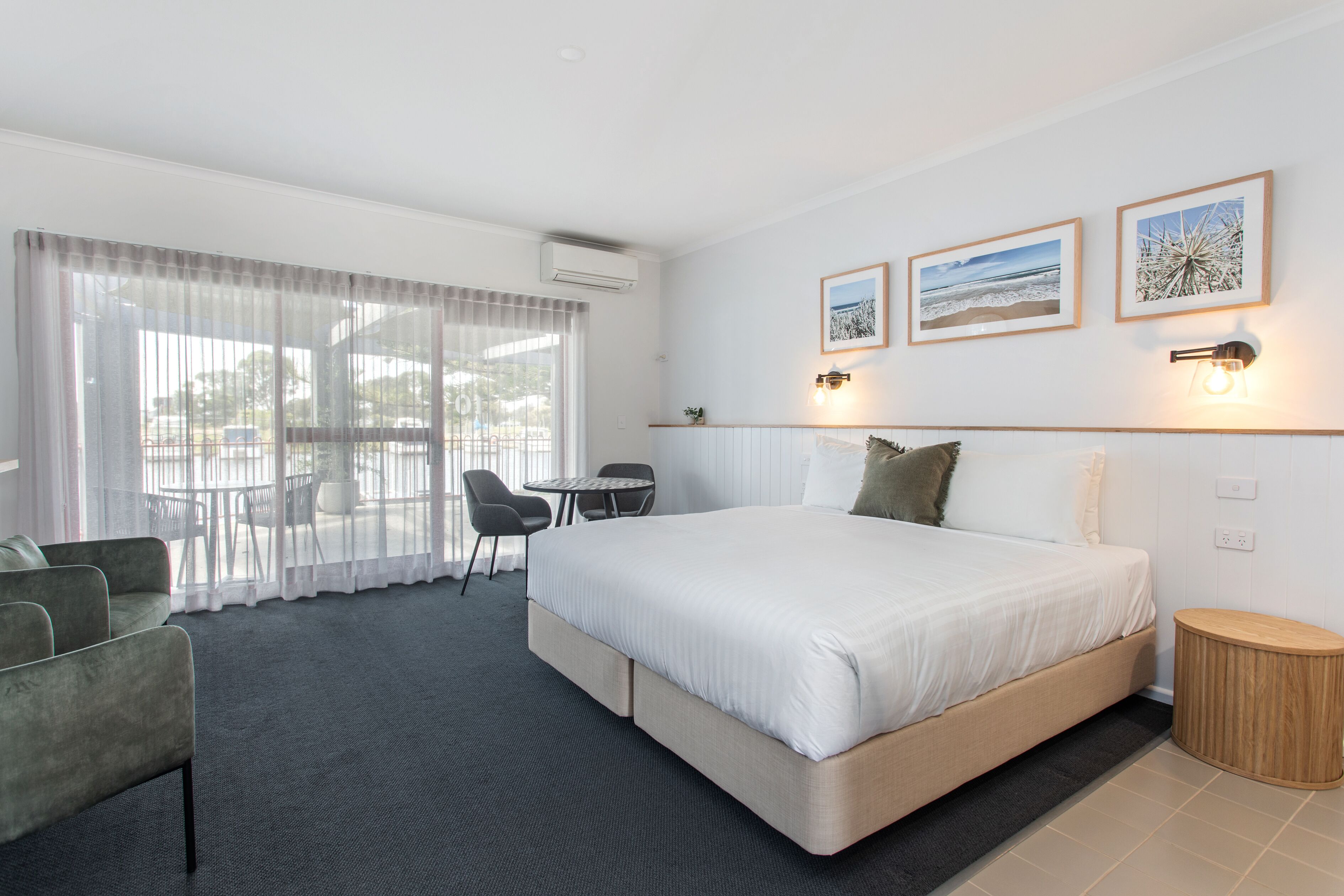 A modern hotel room with a large bed, a seating area with a table and chairs, and a view through large windows.