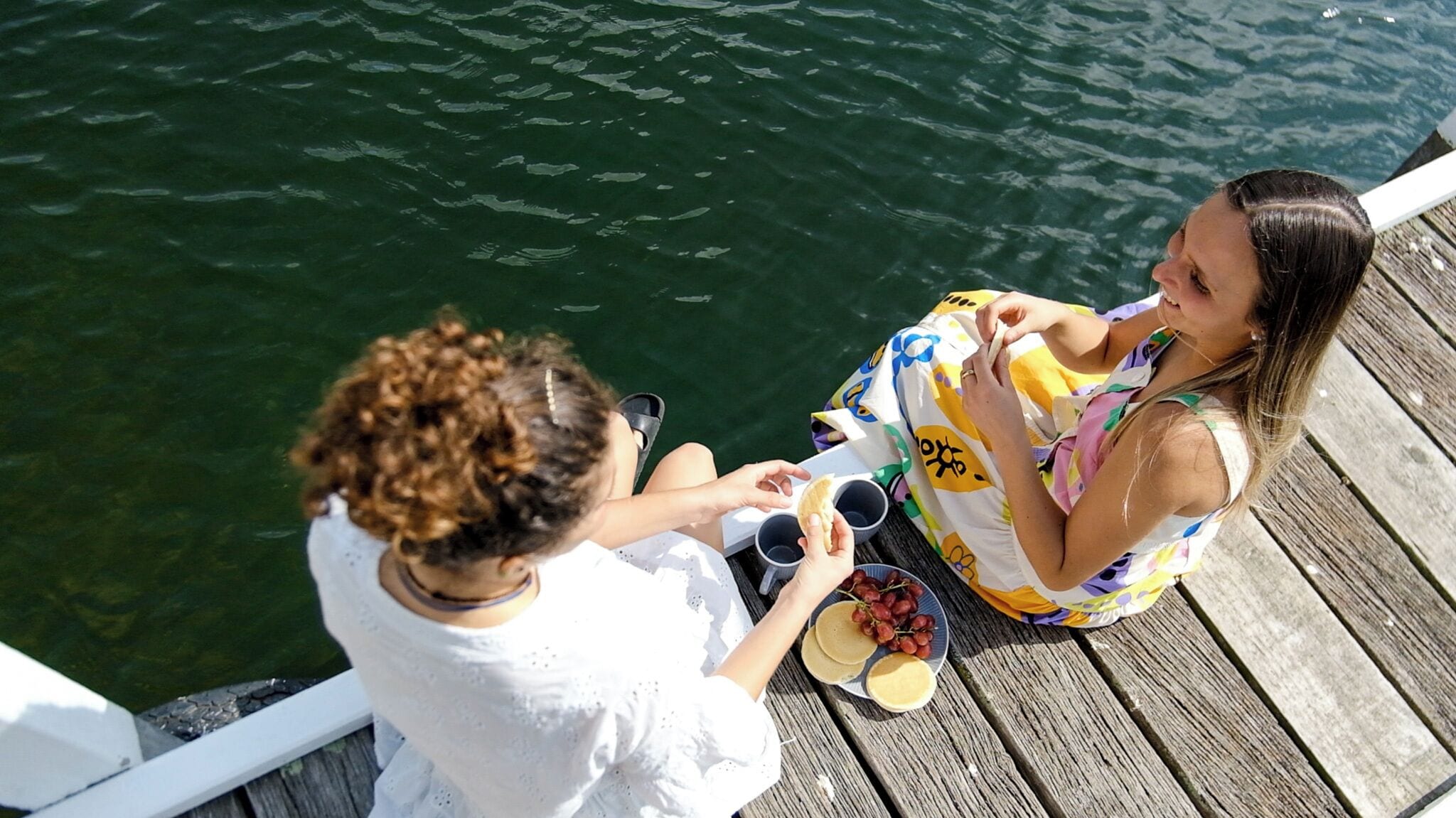 Dos personas sentadas en un muelle de madera junto al agua, sosteniendo bebidas y aperitivos.