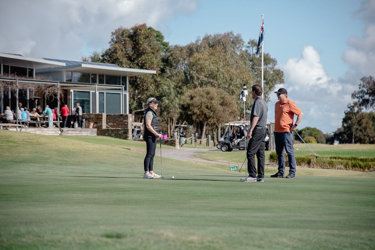 A golf course with a modern clubhouse, a flagpole, and a group of people standing on the green.