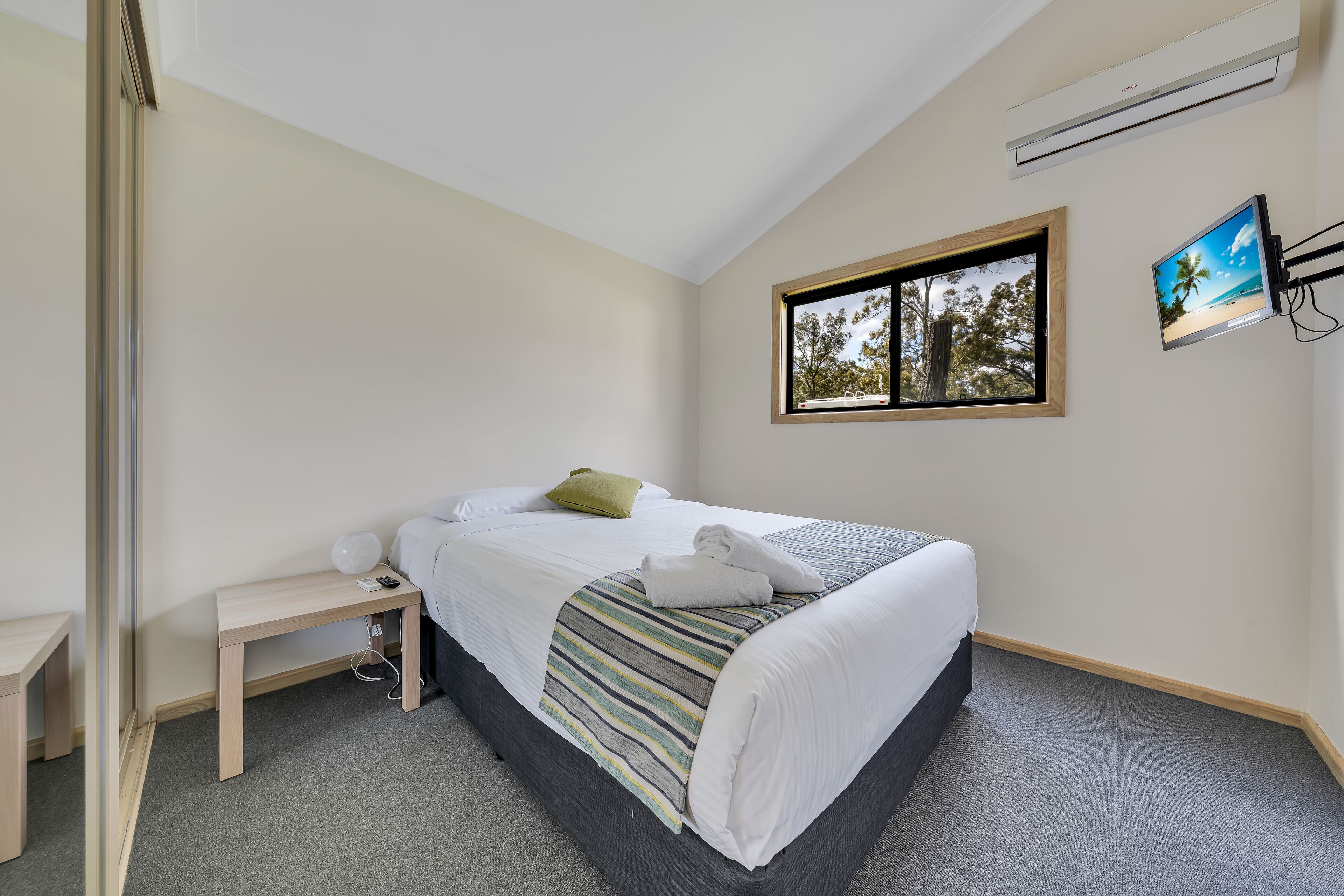 A neatly made bed with a striped blanket, a bedside table with a lamp, and a window with a view of trees.